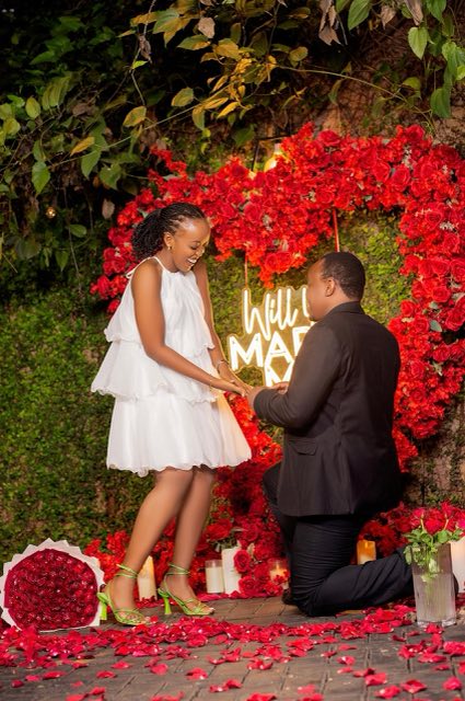 Brian on one knee proposing to Angella in front of a heart of red roses